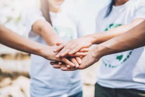 Un grupo de personas hacen equipo colocando sus manos en el centro en señal de unión y compromiso por una causa social. Esta imagen representa la importancia del trabajo en equipo y la colaboración para lograr un cambio positivo en la sociedad. La imagen transmite un mensaje de solidaridad